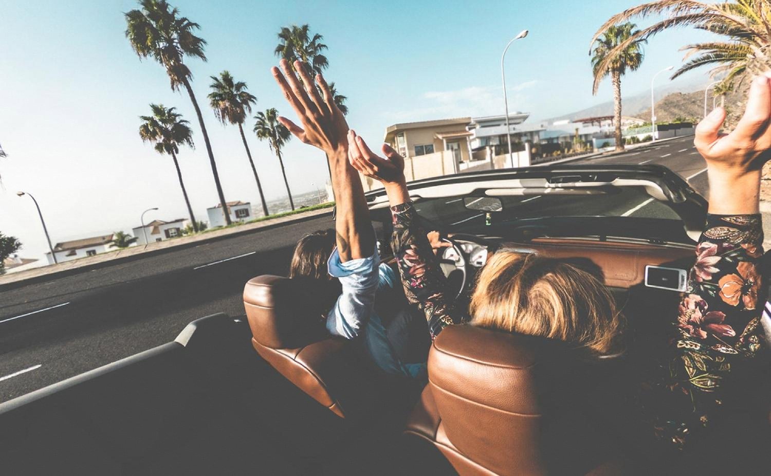 Two travellers with hands in the air riding in a convertible on a palm-lined Australian road